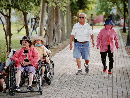 Older adults sitting in wheelchairs and walking on a paved path in a park on a sunny day.