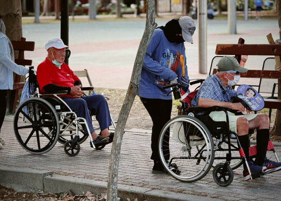 Two elderly men in wheelchairs, wearing face masks, are accompanied by a person standing nearby in a park.
