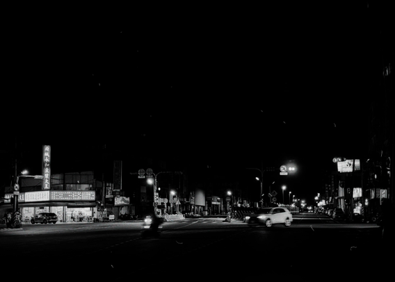 Nighttime city street with illuminated signs, a few cars and a motorbike crossing the intersection under streetlights.