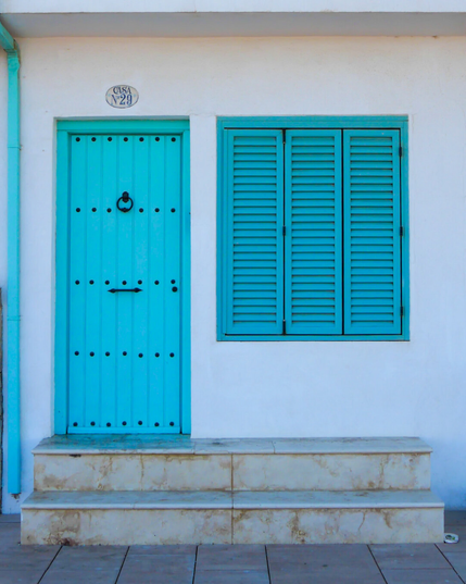 A small white Mediterranean house with a bright turquoise wooden door and matching closed window shutter. The door has  black metal studs, knocker, and handle. A small oval ceramic sign over the door reads Casa N°.29 in a fun font. There are two marble steps in front of the house.