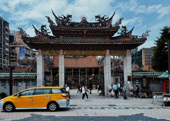 一座传统寺庙的寺门前，有人在散步和交谈，一辆黄色出租车停在前面。