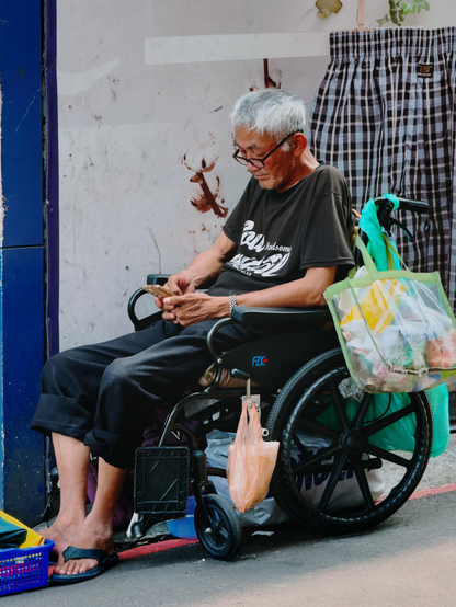 一位老人在轮椅上，穿着黑色T恤和长裤，专注地看手机，背景是墙壁和挂着的布料，旁边有包装袋和垃圾。