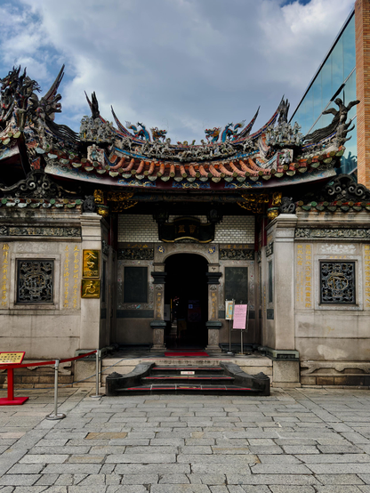 一座具有传统装饰的寺庙建筑，门前有台阶和警示杆，屋顶有龙和凤的雕塑，背景是蓝天白云。