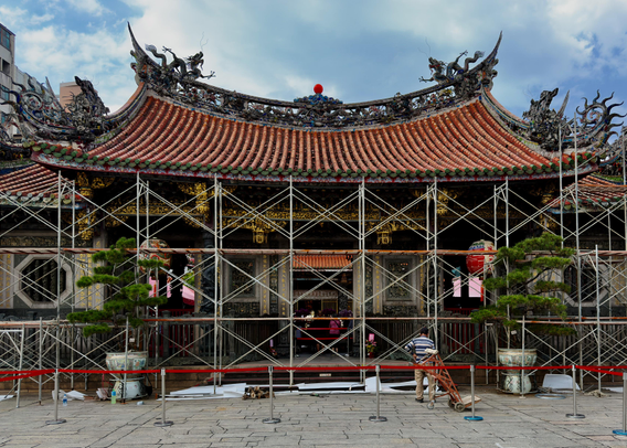 为修缮中的传统寺庙，前景有施工人员和工具，寺庙屋顶尖角装饰复杂，天空多云。
