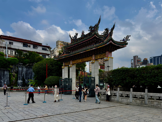 一座传统华丽的牌楼前有人排队，背景是都市建筑和瀑布，天空多云。