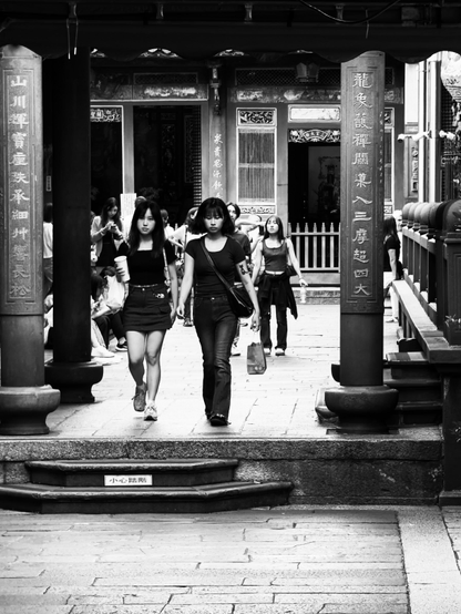 两名年轻女子手牵手走过寺庙入口，背景有其他人休闲聊天或使用手机。