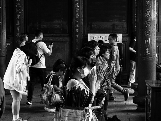 一群人在寺庙里祈祷，部分人戴口罩，有人在弯腰，有人在站立看手机。