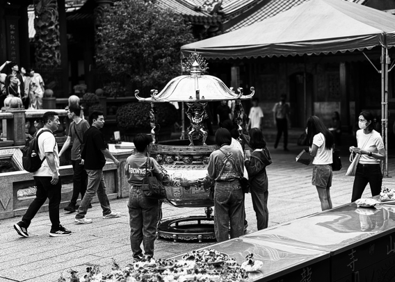 在寺庙前，人们围绕着一个精美的祭祀香炉，有人祈祷，戴口罩，黑白照片充满宗教和文化氛围。
