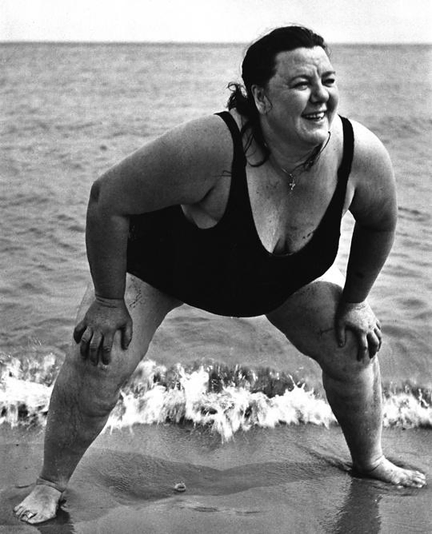 une femme puissante en maillot de bain une pièce, dos à la mer, en position type combat de sumo, prête à affronter le réchauffement climatique