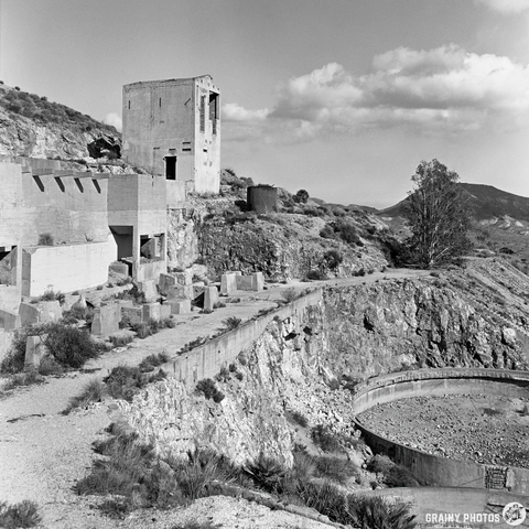 A desolate landscape featuring crumbling concrete structures and rocky terrain under a vast sky with scattered clouds. The remnants of architectural elements hint at a once-thriving site, now abandoned and reclaimed by nature.