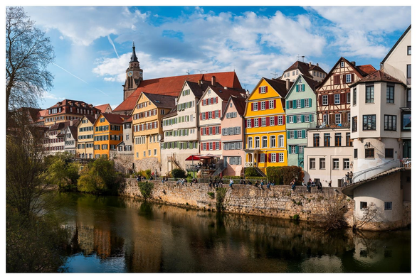 Große bunte (Fachwerk)Häuser stehen dicht nebeneinander an einem Fluss. Im Hintergrund ist auch eine Kirche zu sehen.