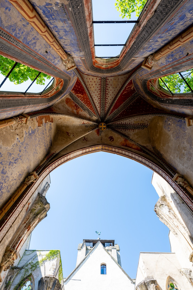Eine vertikale Untersichtaufnahme des ruinierten Innenraums einer gotischen Kirche unter blauem Himmel. Der Fokus liegt auf dem kunstvoll bemalten, noch teilweise erhaltenen Kreuzrippengewölbe mit rotem Sternenmuster und blauen floralen Ornamenten. Die Decke ist teilweise eingestürzt, sodass der Blick durch die Öffnungen direkt in den Himmel und auf die umliegenden Baumwipfel frei ist. Im unteren Bildbereich ist ein Teil des angrenzenden Kirchengebäudes mit einem kleinen Turm und einer Wetterfahne zu sehen.