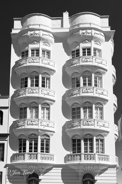 This is a black and white architectural photo in portrait format of an upmarket,  brilliant white, low-rise apartment building located in an upmarket area. Nob Hill, San Francisco (2016).

Dominating most of the image is our almost Gaudi-like apartment building. A hint of neighbouring buildings can be seen to the sides but not enough to identify any detail. In view is the side facade with a tiny bit of the ground floor and four upper floors. What makes this building distinctive are the delicately decorated semi-circular balconies surrounding the three vertical rectangular bay windows. The balcony on the first floor has an ornate low wall or railing at the front divided into three sections with multiple vertical rectangular slots, arched at the top and bottom. The balconies and windows above have an identical shape other than the front of the balconies have elaborately decorated horizontal rectangular, masonry reliefs in three sections surrounding the edge, the two outer pieces being noticeably longer than the centre section. This continues to the upper floor where there is a faux balcony devoid of any detail and low wall surrounding what might be a flat roof. On the right of the building a small amount of further balconies can be seen extending from what is the front facade. To the sides of the building and above is a clear and cloudless sky, rendered an ink black. 