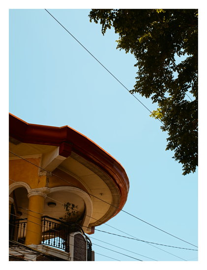 The curved corner of a yellow building with an arched balcony and reddish-orange roof trim. Dark power lines cut diagonally across a clear, bright blue sky, with the dark green leaves of a tree framing the top right corner. - Google Gemini 3.1 Pro Preview