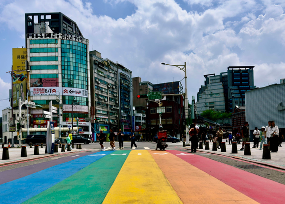 A busy city crosswalk painted in rainbow colors with pedestrians, scooters, and tall buildings under a partly cloudy sky.