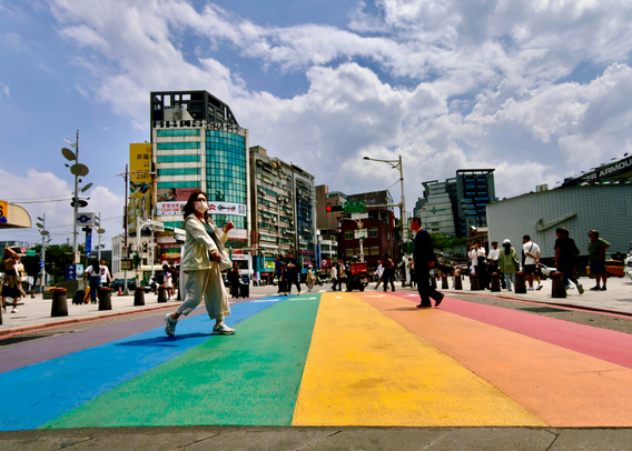A busy city street with people crossing a rainbow-colored crosswalk, high-rise buildings, and a partly cloudy sky above.