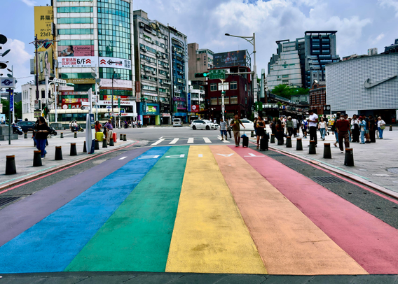 A busy urban street scene with a colorful rainbow crosswalk and pedestrians crossing in a city with tall buildings and advertisements.