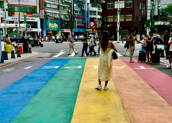 A woman in a floral dress standing on a rainbow-colored crosswalk in a busy city intersection.