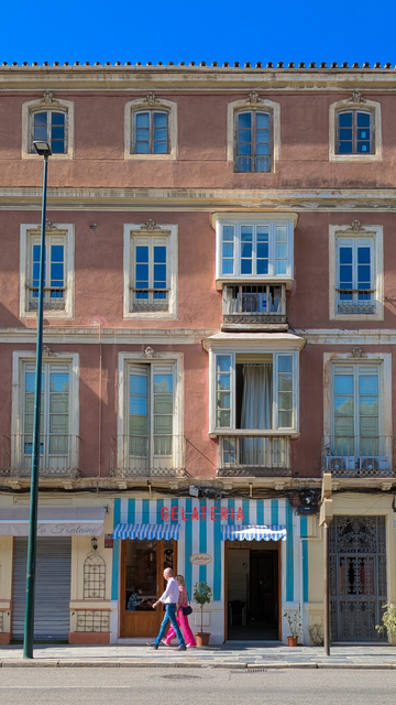 A symmetrical shot of a beautiful building in Málaga, Spain. The pink stucco facade is covered with beautiful windows of different sizes and shapes. The windows reflect the bright blue sky and the greens of the trees across the way. On the ground floor is a gelato shop with a blue and white striped frontage and a GELATERIA sign in pink neon. A couple is walking past, the man is in blue jeans and the woman has on pink trousers which match the blue and pink of the shop perfectly.