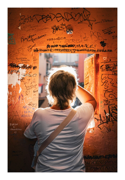 A photograph of a person with short, grey hair and wearing a white t-shirt and a beige shoulder bag. They are standing with their back to the camera, gazing out of the small window in a brightly lit wall covered in colourful graffiti. The sun illuminates her hair, casting a warm glow. The wall is orange, with black and green graffiti. She's taking a photo of the canal.
