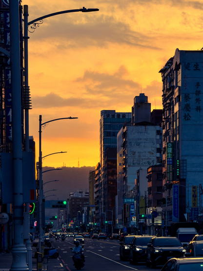 日落时分的繁忙城市街道，道路两旁有许多车辆和行人，背景是高楼林立的城市天际线。