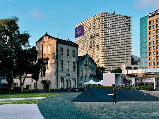 A person standing on a paved area with modern and old buildings in the background under a clear blue sky.