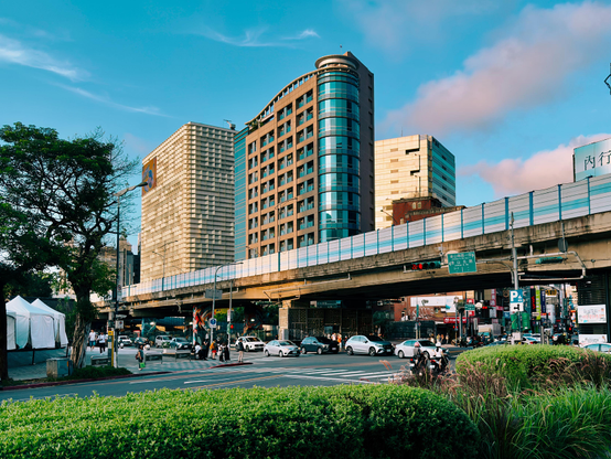 Urban street scene with tall buildings, a bridge, cars, and pedestrians, under a partly cloudy sky.
