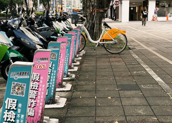 A row of parked scooters and a yellow bike on a city sidewalk, with a tree and pedestrians in the background.