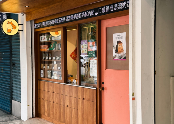 A small bakery storefront with wooden accents, glass display showing packaged baked goods, pink door with poster, and illuminated sign with a cartoon girl drinking.