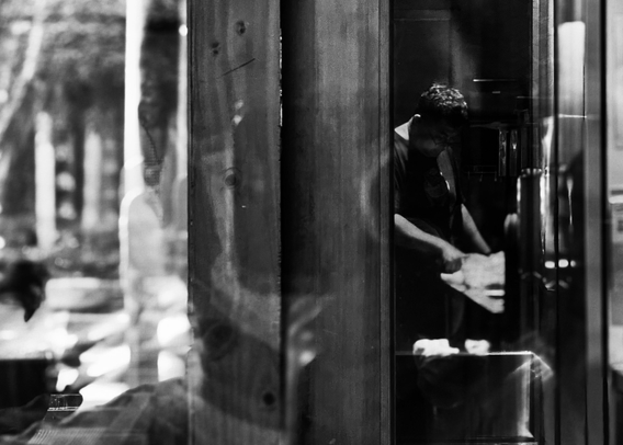 A person is sitting inside a small shop or booth, possibly reading or writing, with reflections of people outside visible on the glass.
