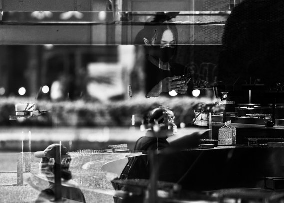 A person wearing a mask looks at their phone through a shop window, with reflections showing street scenes and other individuals inside.