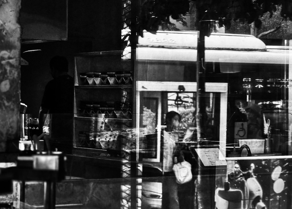 Reflections of people inside a store, with a bicycle and shop items visible through the glass window, in black and white.