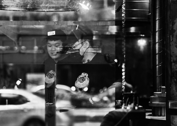 Reflection of a woman with short hair and a face mask on a glass window in an urban setting at night.