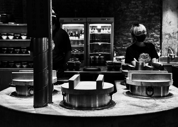 A black and white photo of two people wearing masks in a restaurant kitchen, with steaming pots in the foreground.
