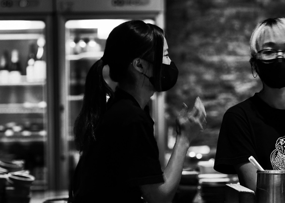 A woman wearing a face mask and glasses, holding a utensil, in a black and white photo with another person beside her.