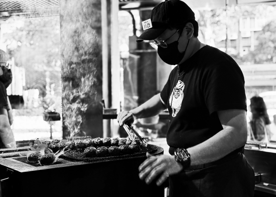 Man grilling meat skewers at a street food stall, wearing a mask, cap, and glasses, with steam rising from the food.