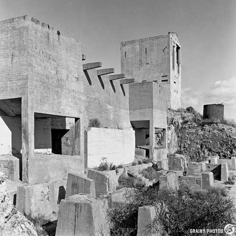A black and white photograph of rugged concrete structures on rocky terrain, featuring angular lines and geometric shapes, set against a clear sky with scattered clouds. The scene evokes a sense of desolation and industrial decay.
