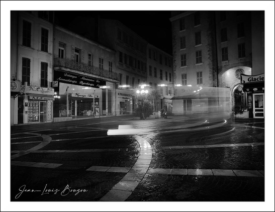 A nighttime city square or broad intersection, rendered in moody black and white, fills the frame with a sense of quiet motion and urban stillness. The buildings rise on either side in tall, dark blocks, their facades lined with windows and shopfronts that glow faintly against the surrounding shadows. Light spills from a few streetlamps and storefront signs, creating bright pools on the pavement and catching the edges of the architecture.
The center of the scene feels almost dreamlike: a long-exposure blur of a passing vehicle arcs through the middle, leaving behind a streak of light that curves across the dark street like a ribbon. The wet ground reflects the lamps and windows, giving the whole image a glossy, mirror-like sheen. The pavement markings in the foreground lead the eye inward, adding structure to the otherwise fluid blur of the scene.
There’s a strong contrast between stillness and movement here. The buildings remain solid and silent, while the light trail suggests life rushing through the night just out of reach. The overall atmosphere is cinematic, slightly lonely, and contemplative—an empty city moment captured with elegance and a touch of mystery.

