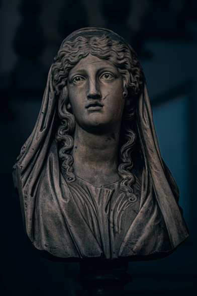 A photograph of a classical stone bust of a woman with long, wavy hair that is partially covered by a draped veil.  The bust is detailed, with textured fabric and smooth facial features. The background is blurred, dark, and indistinct (she's at the stairs of one of the university buildings) The colour palette is cold and blue and moody.
