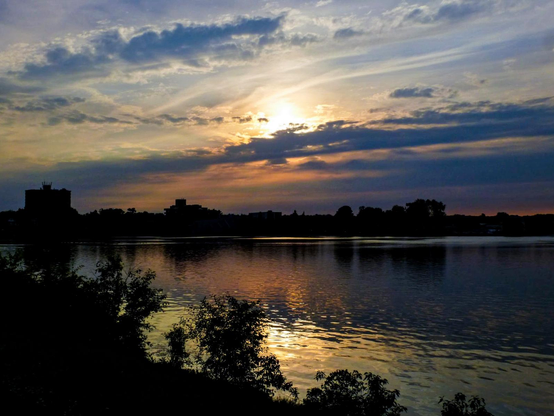 The warm glow of sunset reflects off the calm water of a lake. The sun is partially hidden by clouds and the silhouette of the distant shoreline and the bushes in the foreground are seen.