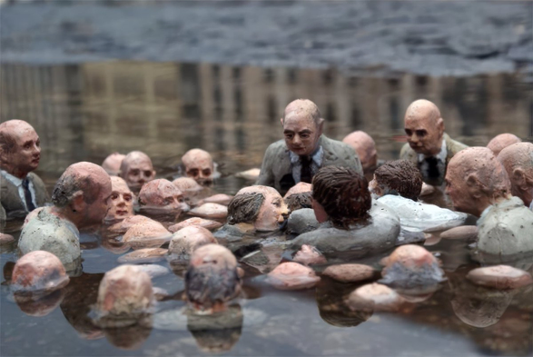 A close-up photograph of an art installation featuring miniature sculpted busts of men in business suits. The figures are placed in a puddle, appearing partially submerged with the water reaching their chests or necks.