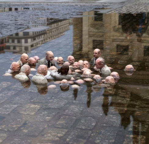 A wider photograph of the same street puddle installation. The miniature sculpted men in suits are clustered in the water, and the wet surface of the puddle reflects a nearby multi-story building.