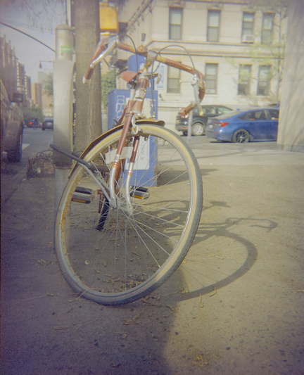 A rusty steel road bike with a flat tire seen from the front, casting a shadow on the sidewalk