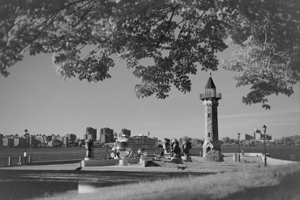 Looking beneath a tree, at the Roosevelt Island lighthouse and a public art project with many sculptures of human faces and shiny metal spheres