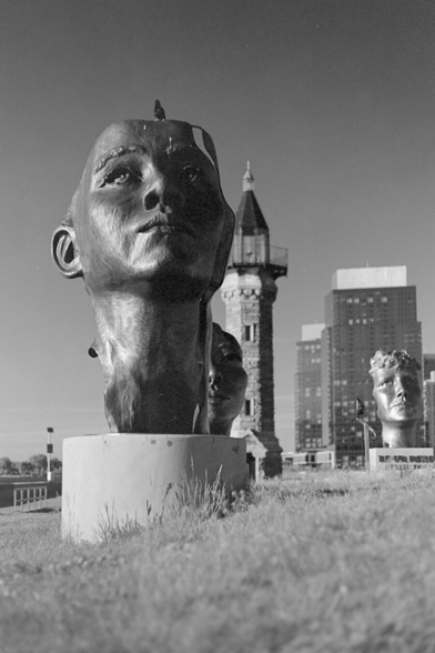 A giant sculpture showing a human face, in harsh sun, on top of grass that looks bright with Wood effect. The lighthouse is in the background.