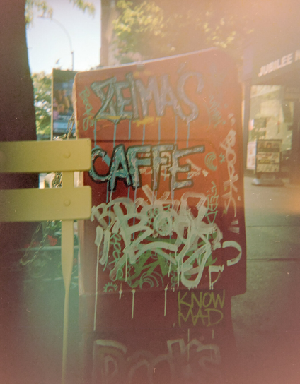 A red newspaper box with garish graffiti, by a yellow metal chair, in the shade
