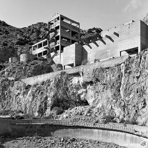 A black-and-white image of a rugged hillside with a partially constructed concrete building. Surrounding rocky terrain and a curved structure in the foreground suggest an abandoned site, emphasizing a desolate and industrial atmosphere.