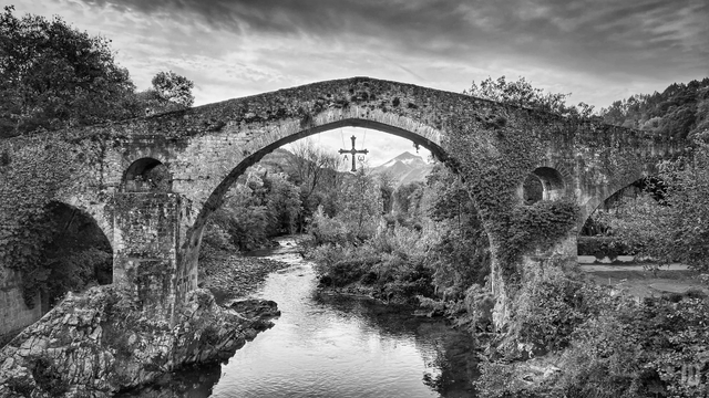 La imagen muestra un puente de piedra antiguo con un gran arco central que cruza un río rodeado de vegetación frondosa. En el centro del arco cuelga una cruz con las letras “A” y “Ω”. Al fondo se observan montañas y un cielo parcialmente nublado, creando un paisaje natural y sereno.
