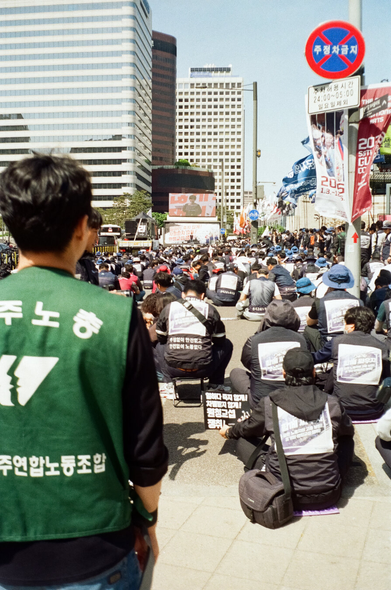 민주노총 결의대회가 열리는 서울 도심. 멀리 대형스크린 앞에 조합원들이 앉거나 깃발을 들고 있다. 카메라 근처에는 민주연합노조 초록색 조끼를 입은 조합원 등 몇 명이 서서 스크린을 바라보고 있다. 화면 뒤로는 거대한 마천루가 늘어섰다.