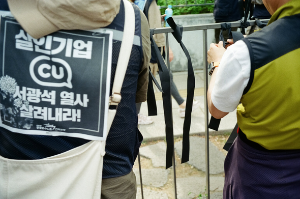 청와대 앞 도로 난간에 민주노총 조합원들이 검은 리본을 묶고 있다. "살인기업 CU 서광석 열사 살려내라!"는 등자보를 입은 공공운수노조 조합원과 연두색 조끼를 입은 조합원 사이로 검은 리본이 묶여있는 것이 보인다.